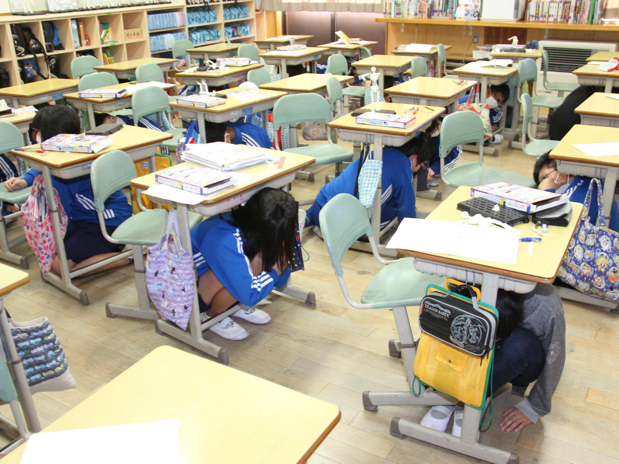 避難訓練で地震発生の校内放送の後、児童が机の下に隠れる様子